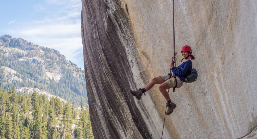 A person wearing safety gear is suspended by ropes mid air against the backdrop of a large smooth rock wall. There are evergreen trees, a mountain and blue skies in the background. 